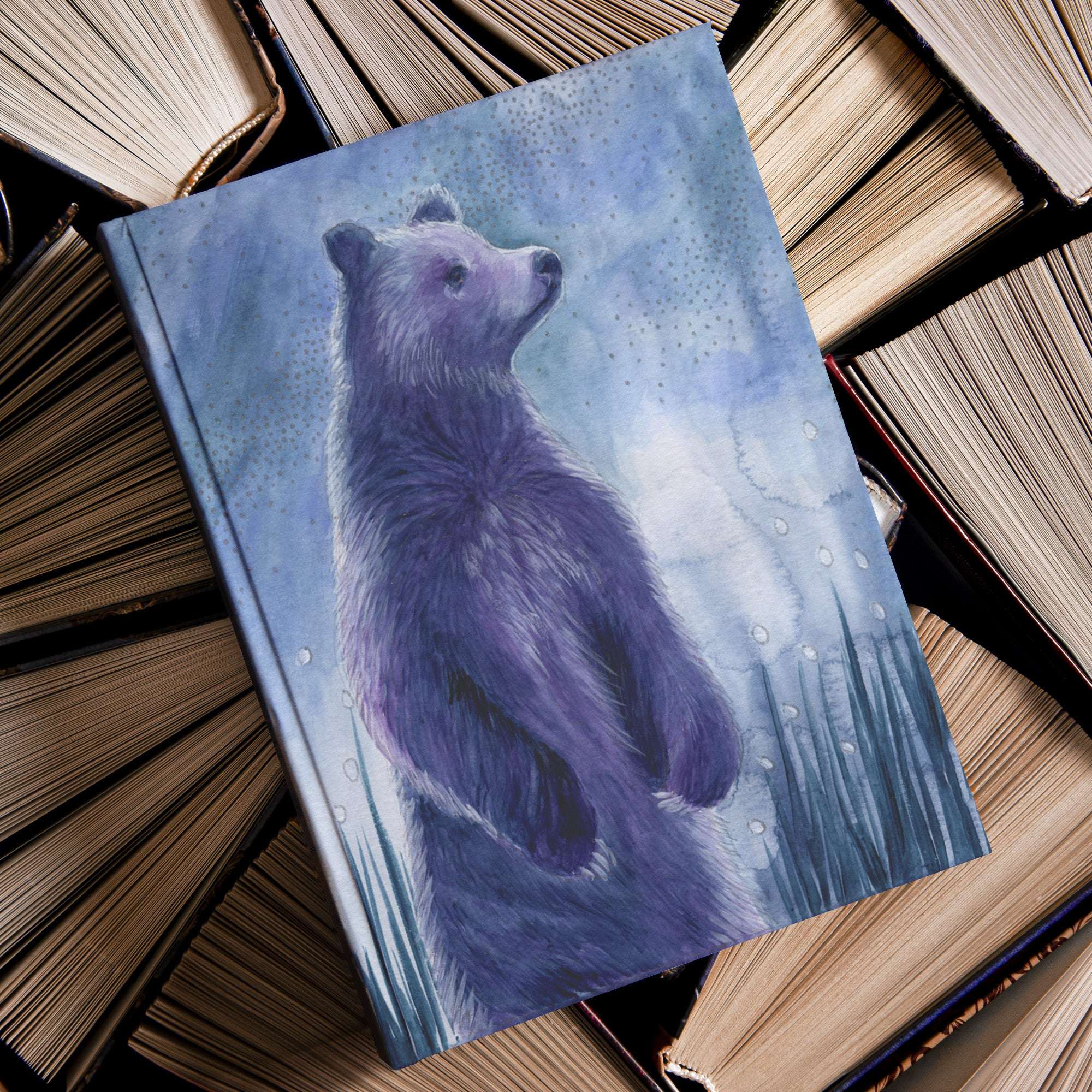 A watercolor painting of a purple bear on the Celestial Bear Journal cover, surrounded by stacks of old books.