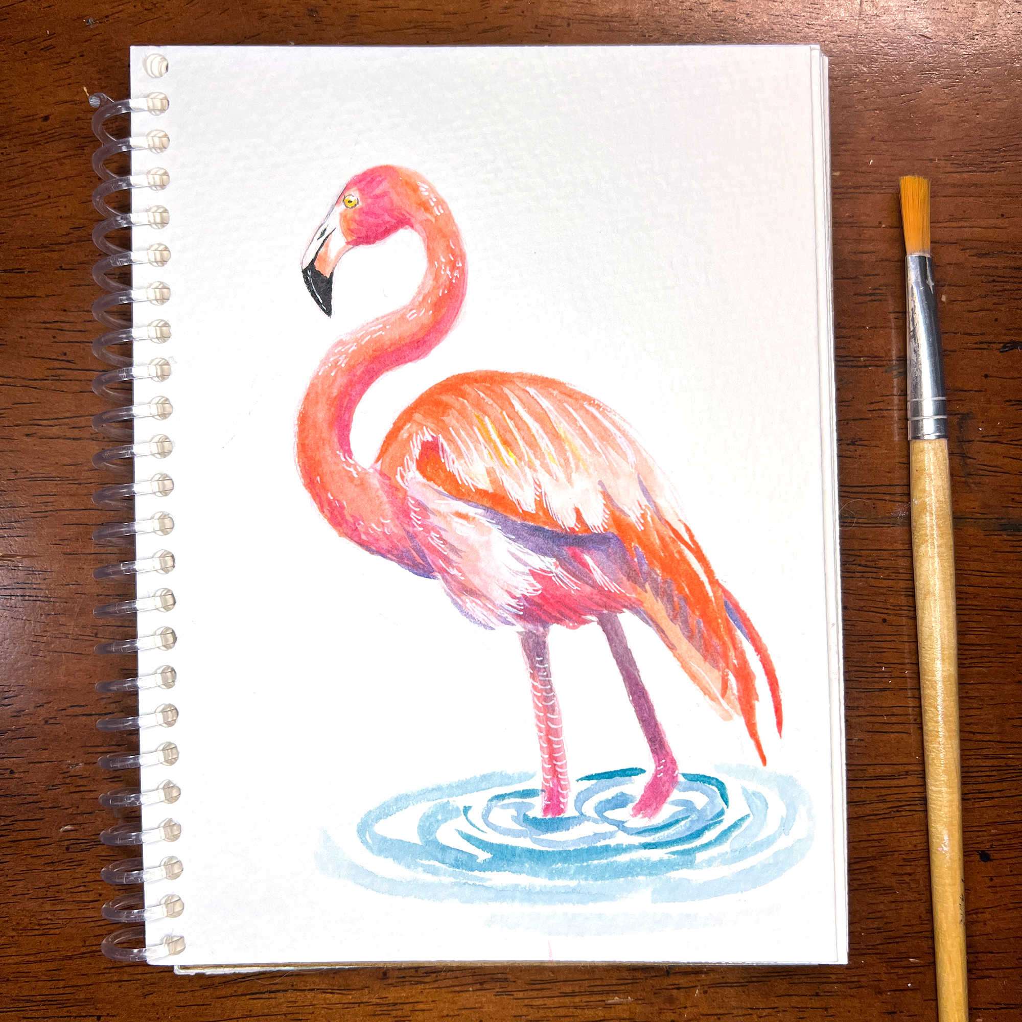 Flamingo watercolor painting on a sketch pad with a brush beside it.