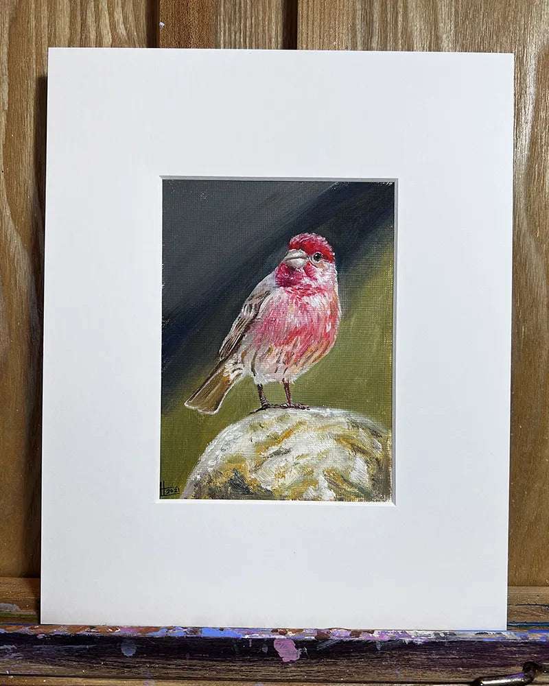 Oil painting of a common rosefinch framed with a white mat on a wooden easel.