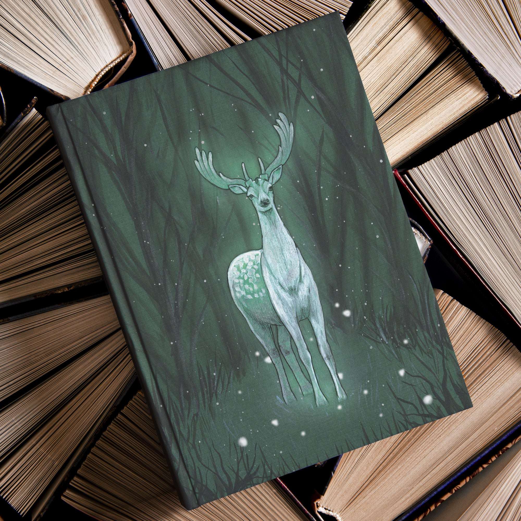 A Enchanted Deer Journal with an illustration of a mystical deer spirit in the forest lies on top of a pile of aged, hardcover books.