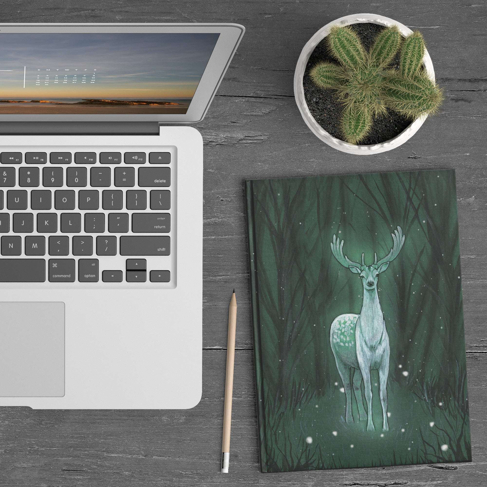 An Enchanted Deer Journal, on a wooden desk, with a painting of a green deer spirit on the cover.