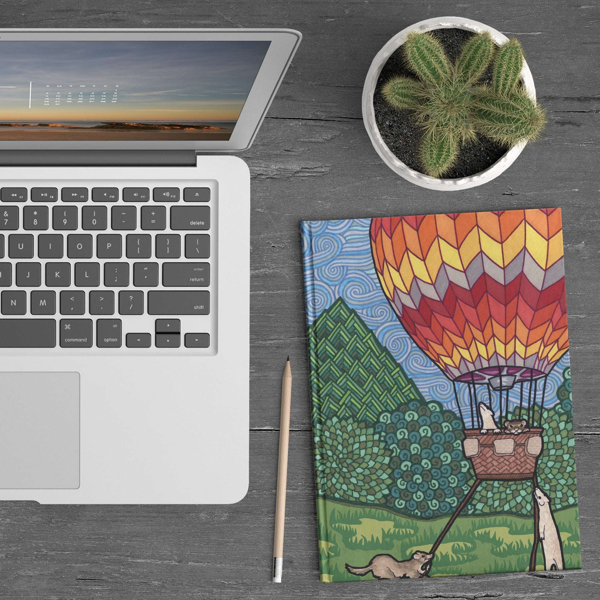 A laptop, closed Ferret Flight Journal with a hot air balloon ferret design, pencil, and a potted cactus on a wooden table.