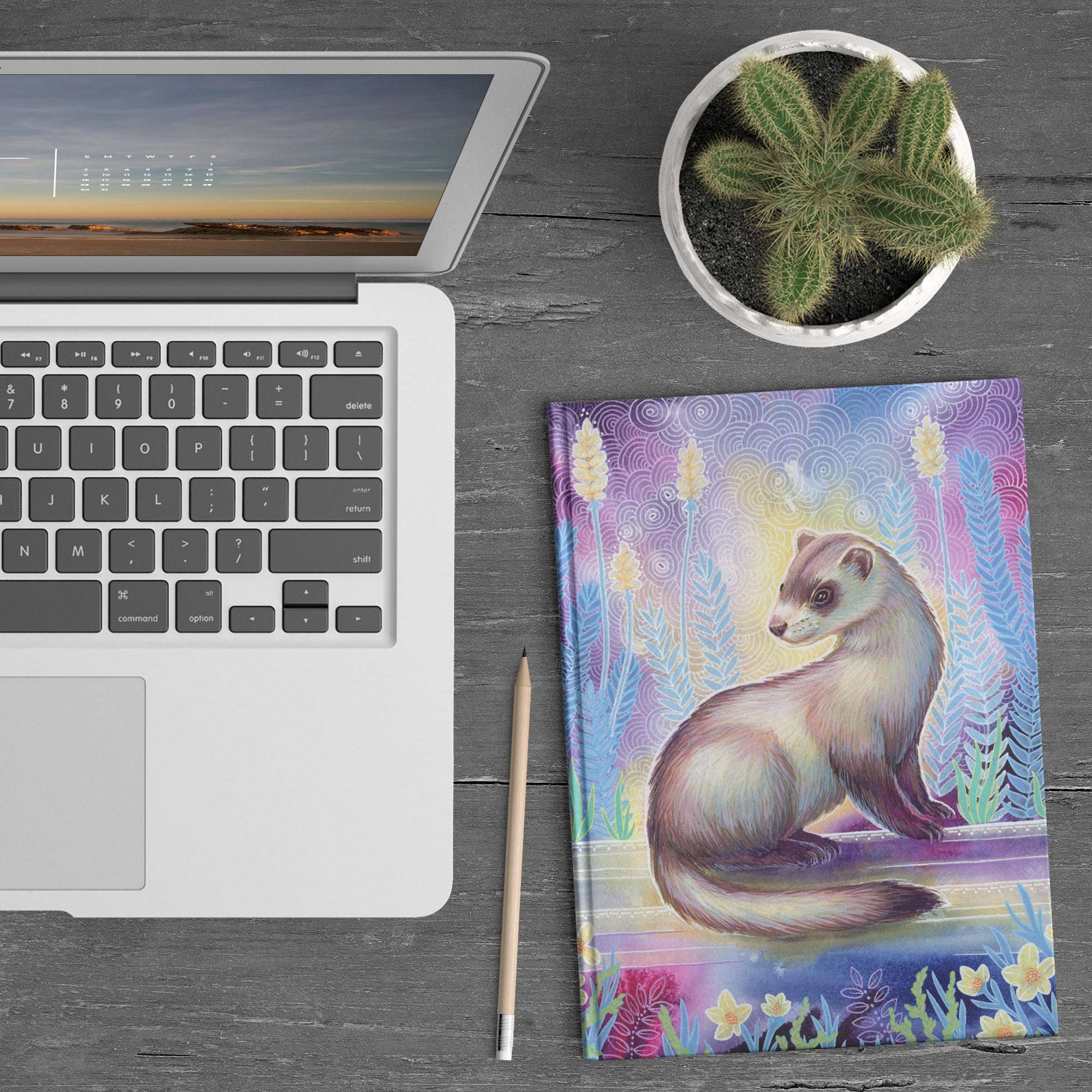 A laptop, Ferret Journal with a colorful ferret illustration, a pencil, and a potted cactus on a wooden table.