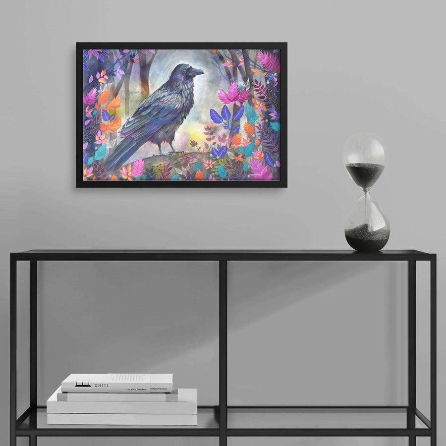 A framed Raven Art Print of a black raven among vibrant flowers displayed on a wall above a modern shelf with books and an hourglass.