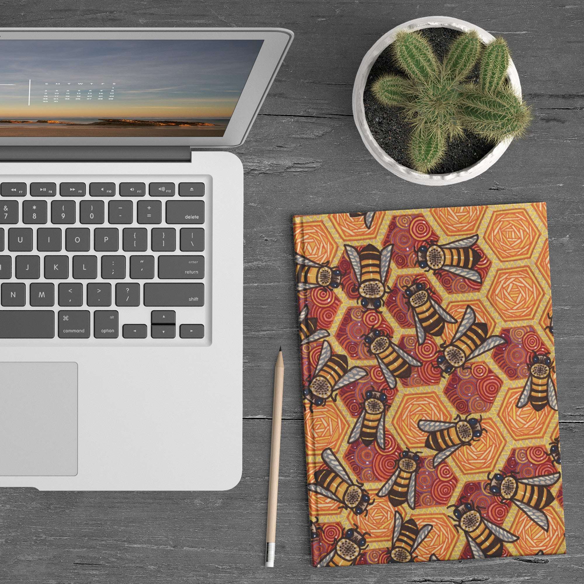 Top view of a workspace featuring an open laptop with a sunset screen wallpaper, a Honeycomb Harmony Journal, a pencil, and a small potted cactus on a wooden desk.