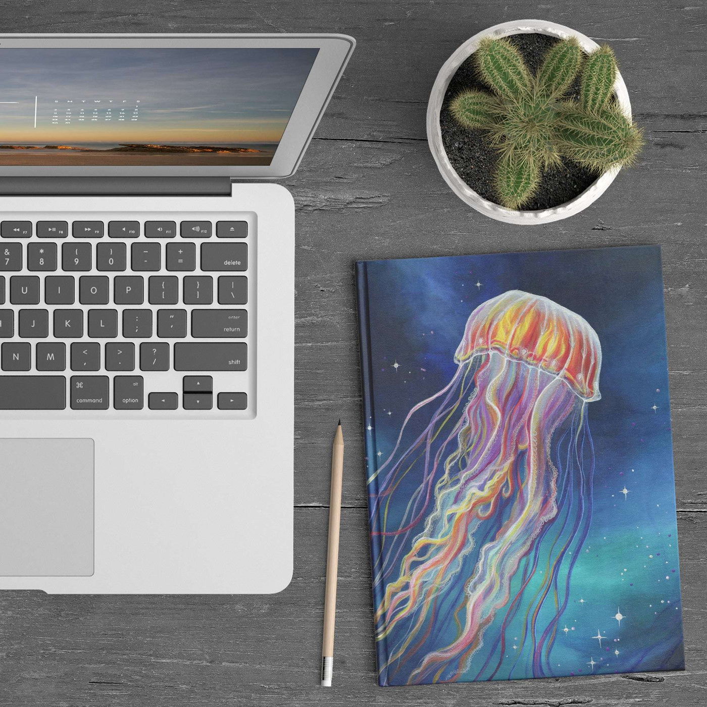 A laptop, Jellyfish Journal, pencil, and a potted cactus on a wooden table.
