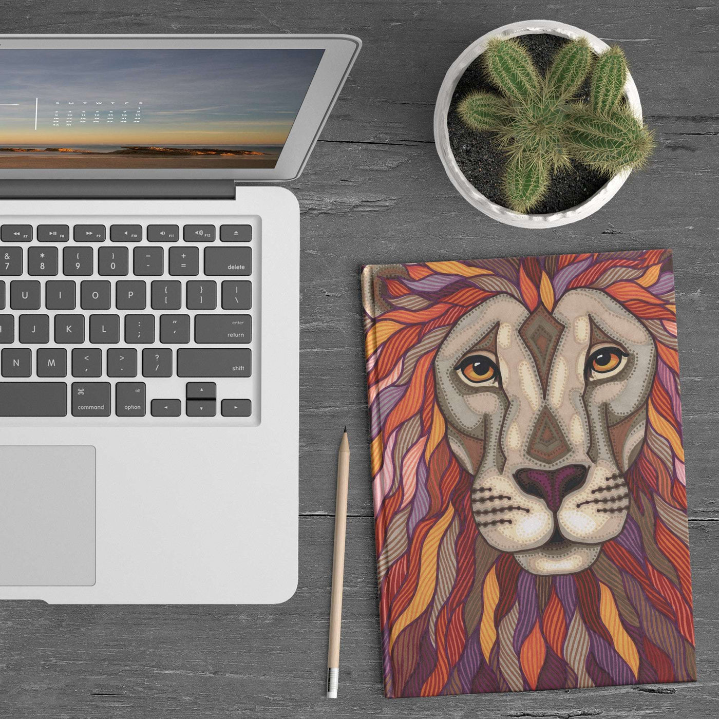 A laptop, Lion Pride Journal, pencil, and small potted cactus on a wooden desk.
