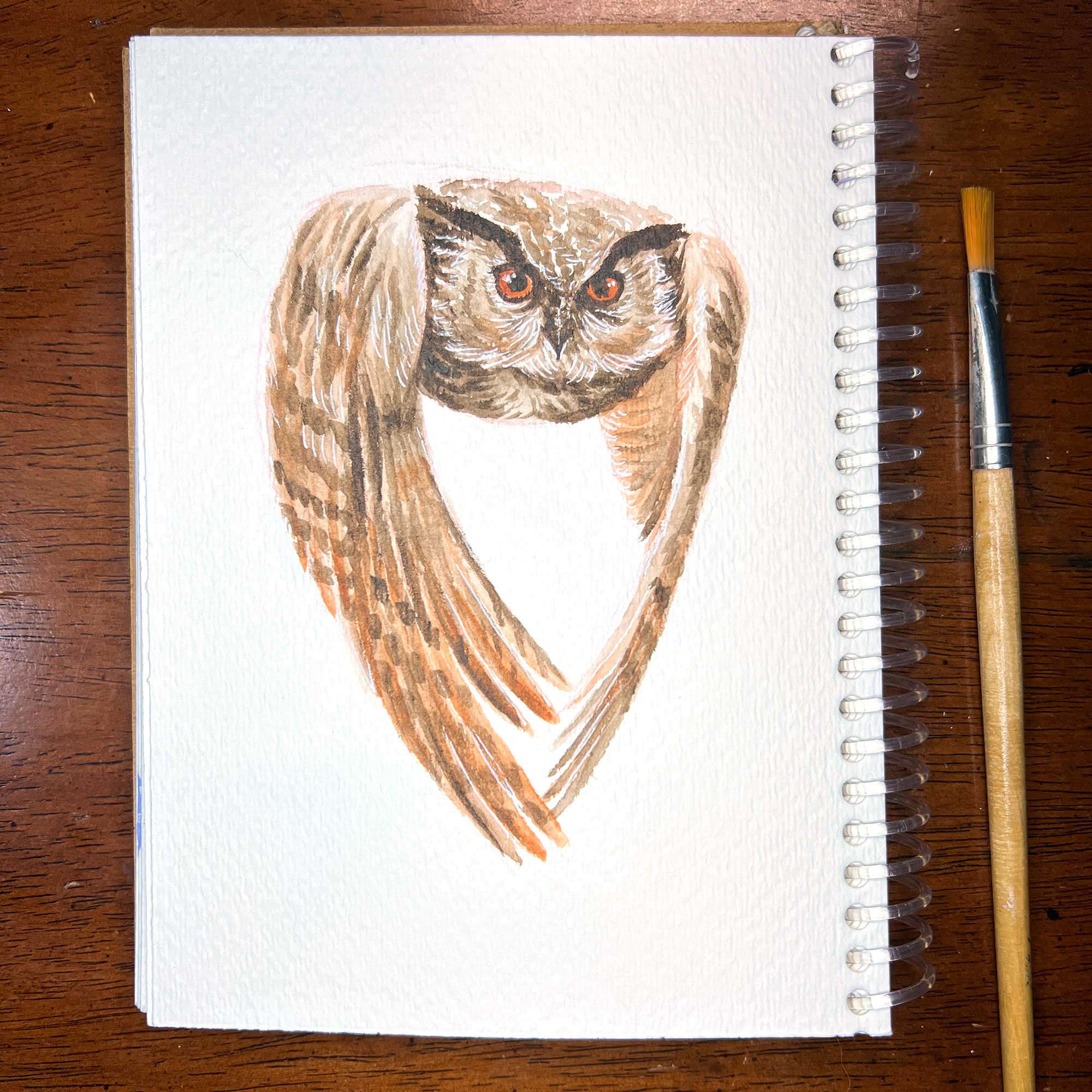 Owl/Rooster - Original Watercolor Painting of an owl on a spiral notebook, with a paintbrush resting beside it on a wooden table.