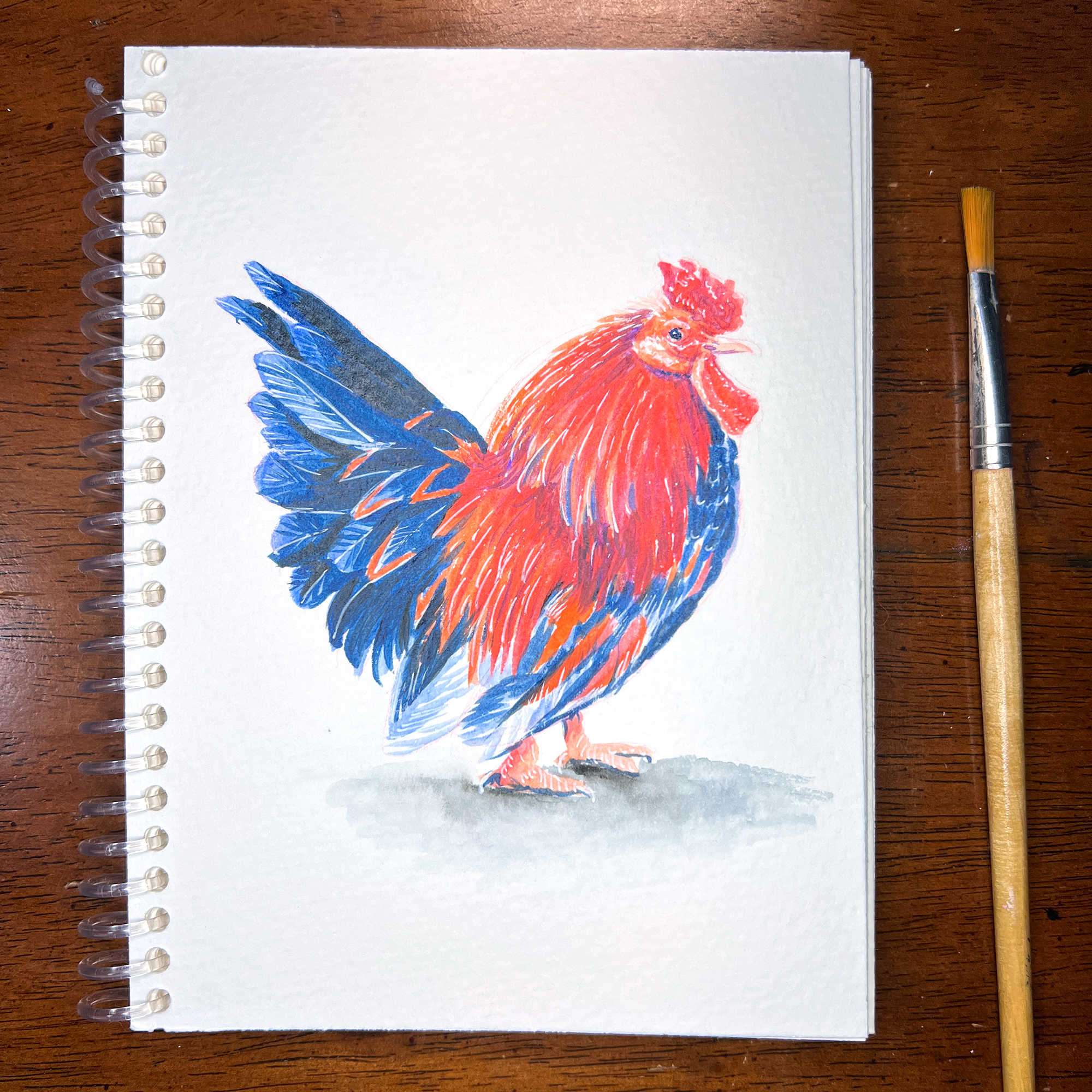 A colorful Owl/Rooster - Original Watercolor Painting on a sketchpad, with a paintbrush placed next to it on a wooden surface.