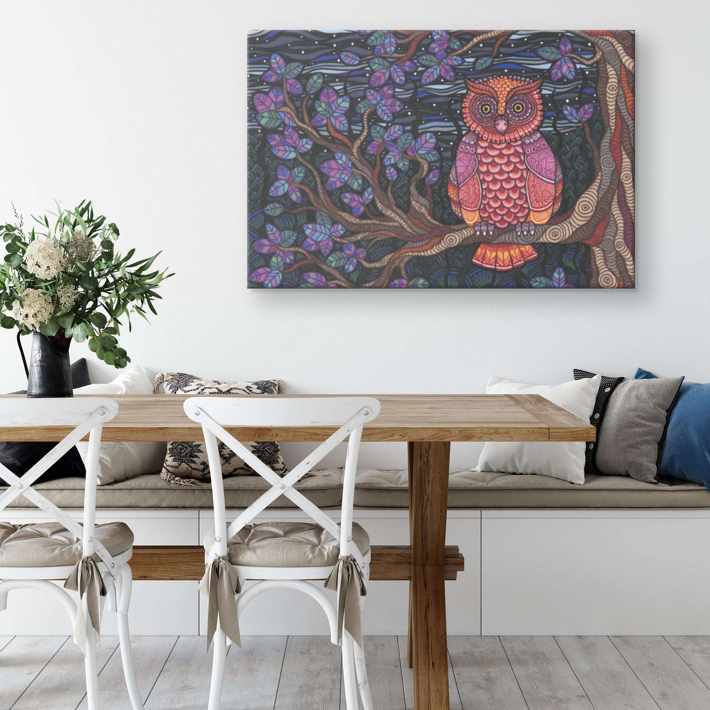 An Owl Tree Canvas Print hung above a dining table with white chairs and a vase of flowers, inside a neatly decorated room.