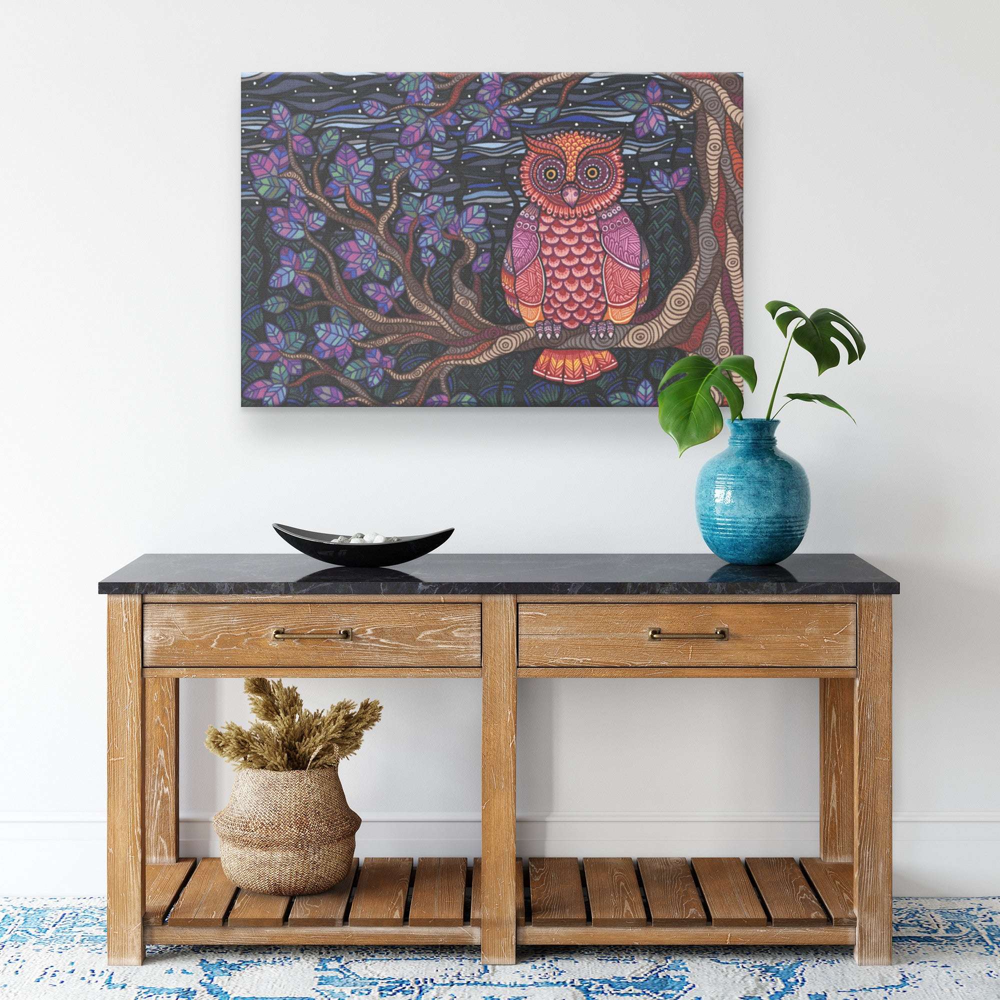 A serene entryway featuring a wooden console table with a Owl Tree Canvas Print above and decorative items including a blue vase and dried plants.