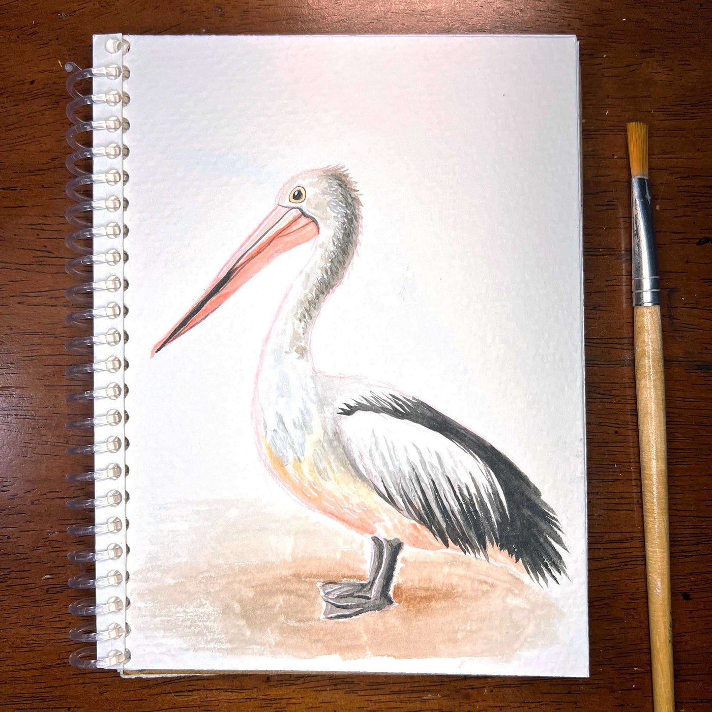 Pelican - Original Watercolor Painting of a pelican on a sketchbook, lying next to a paintbrush on a wooden surface.