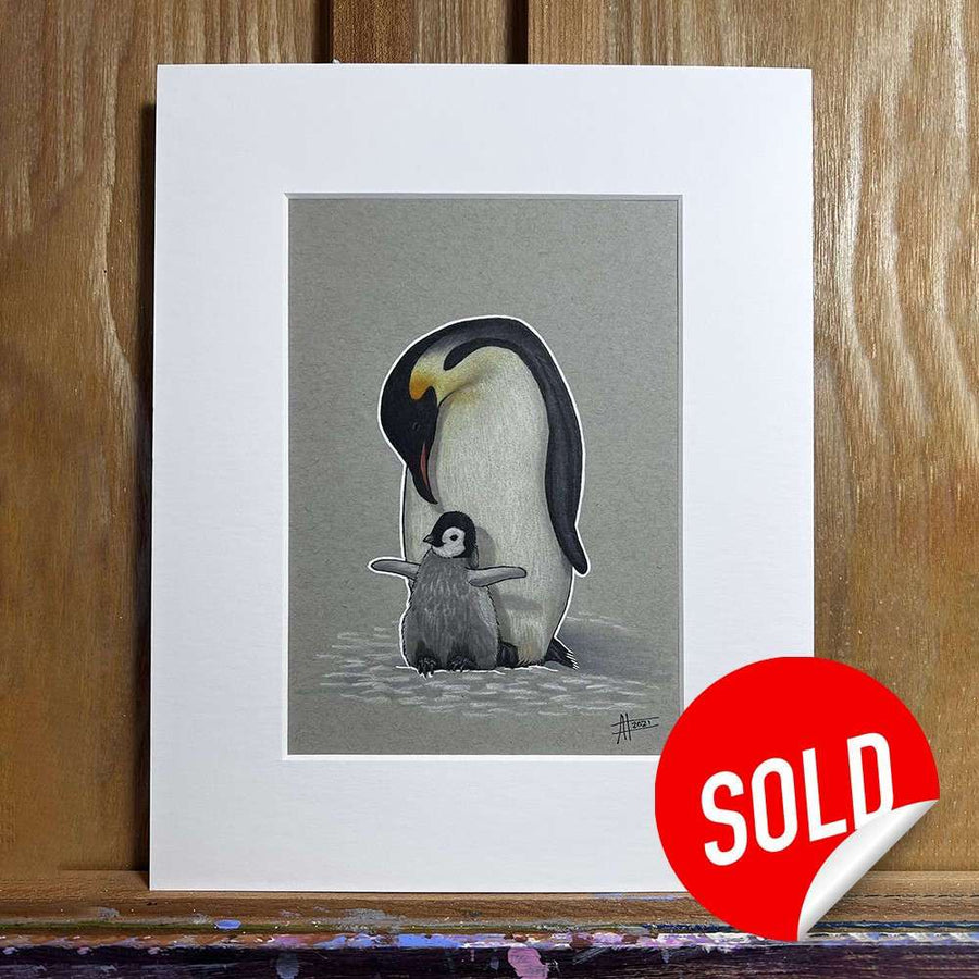 Illustration of a Penguin With Baby - Original Marker Painting with its wing around a chick, displayed with a "sold" sticker on the frame, set against a wooden background.