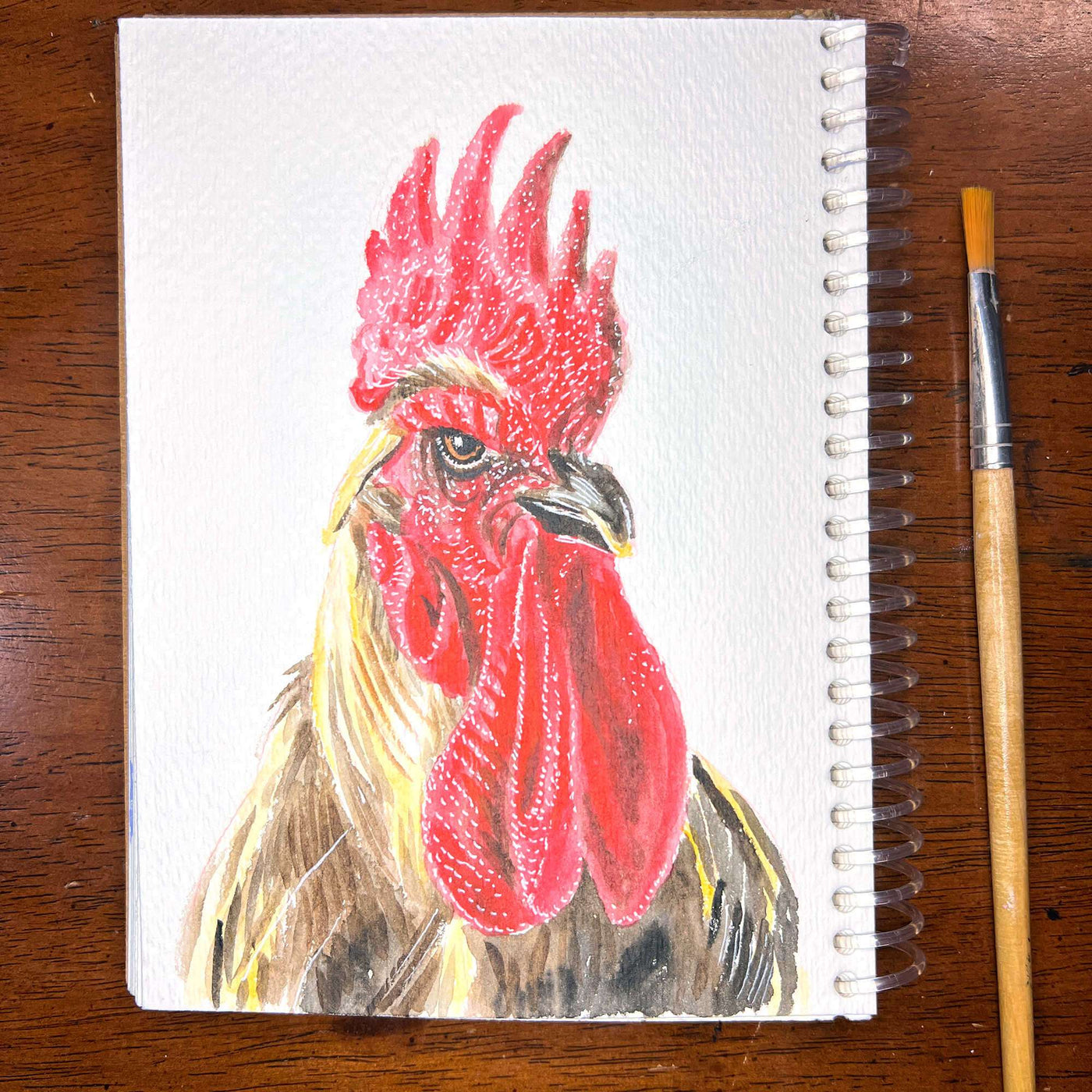 Pigeon/Rooster - Original Watercolor Painting of a rooster on a spiral notebook, with a paintbrush on the right, displayed on a wooden surface.