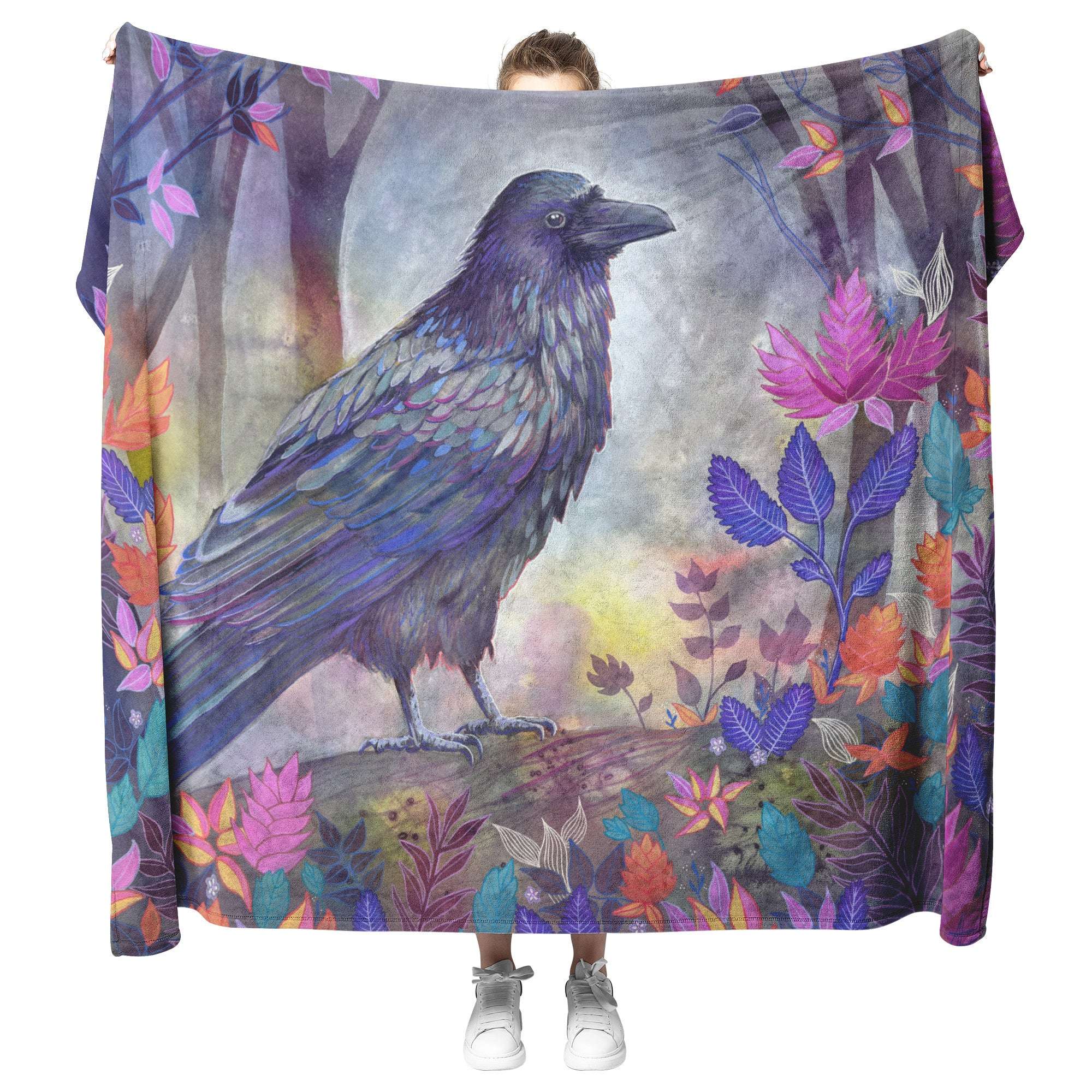 A person holding a Raven Blanket featuring a vivid illustration of a raven in a forest surrounded by colorful leaves and flowers.