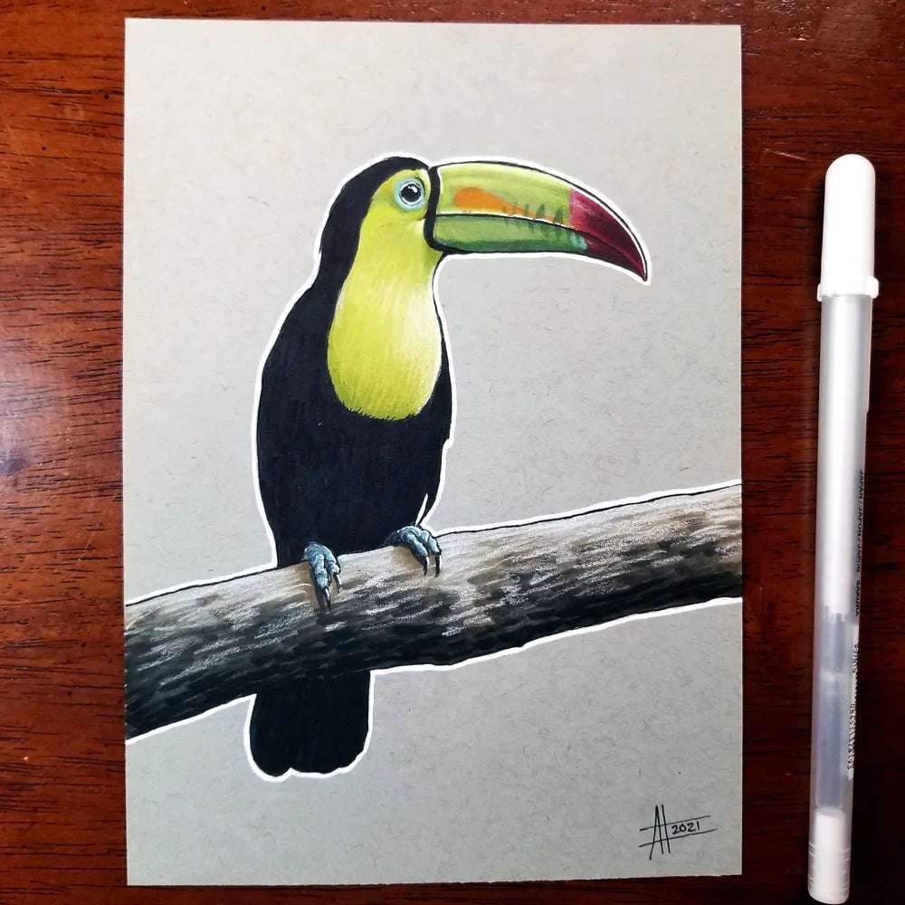Illustration of a colorful Toucan - Original Marker Painting by Amanda Lanford sitting on a branch, with a white pen placed beside the paper on a wooden surface.