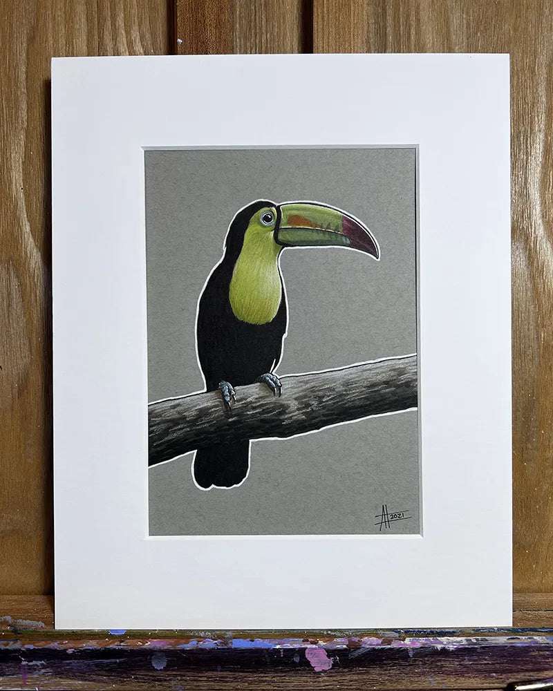A full view of a pen and marker painting of a Toucan Amanda Lanford. perched on a branch, displayed within a white mat and wooden frame against a wooden easel background.