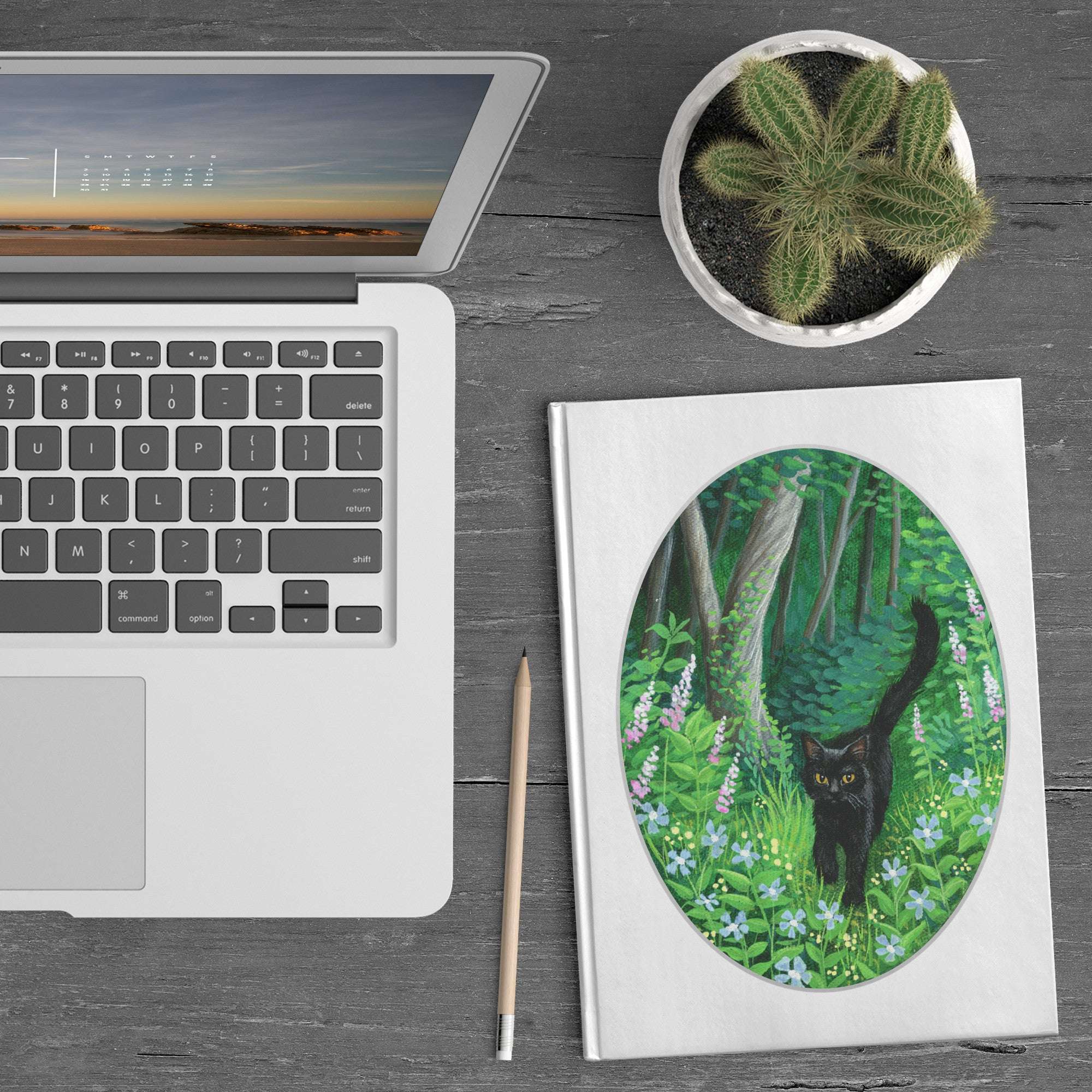 A laptop, Whiskered Welcome Journal with a black cat illustration, pencil, and a potted cactus on a wooden desk.