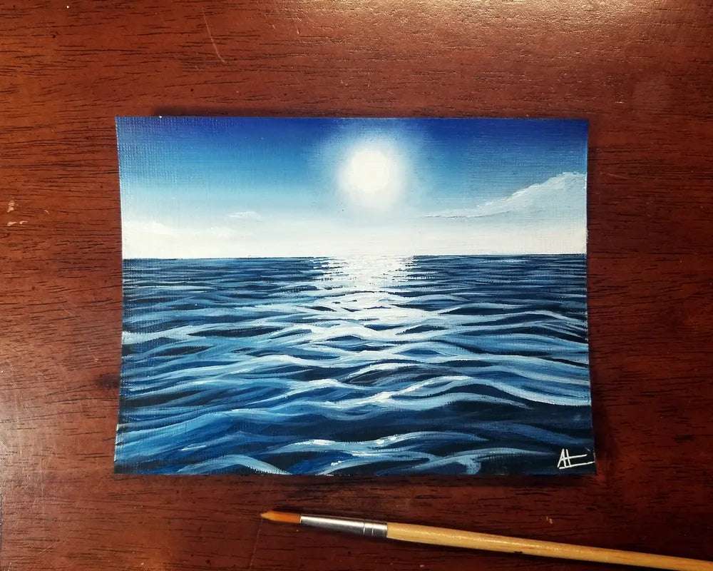 A painting of a Tiny Seascape under a bright moon. the canvas is placed on a wooden surface alongside a paintbrush.