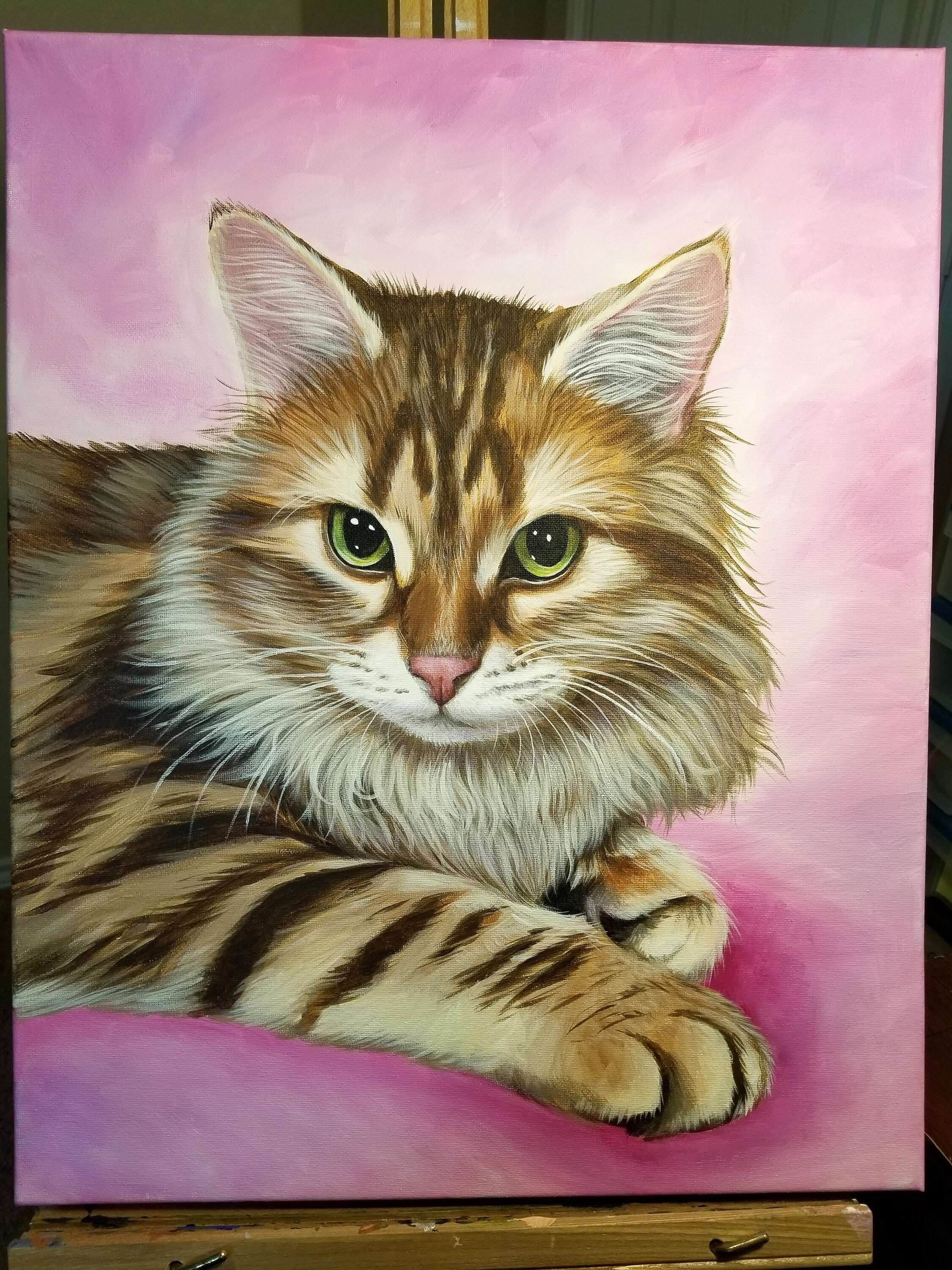 Lifelike pet portrait painting of a tabby cat with green eyes against a pink background, displayed on an easel.