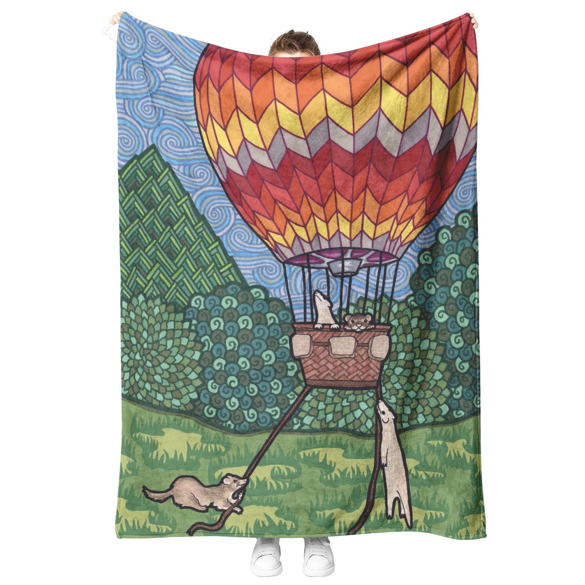A person's holding up a Ferret Blanket, with whimsical designs featuring ferrets in a hot air balloon and stylized landscape.