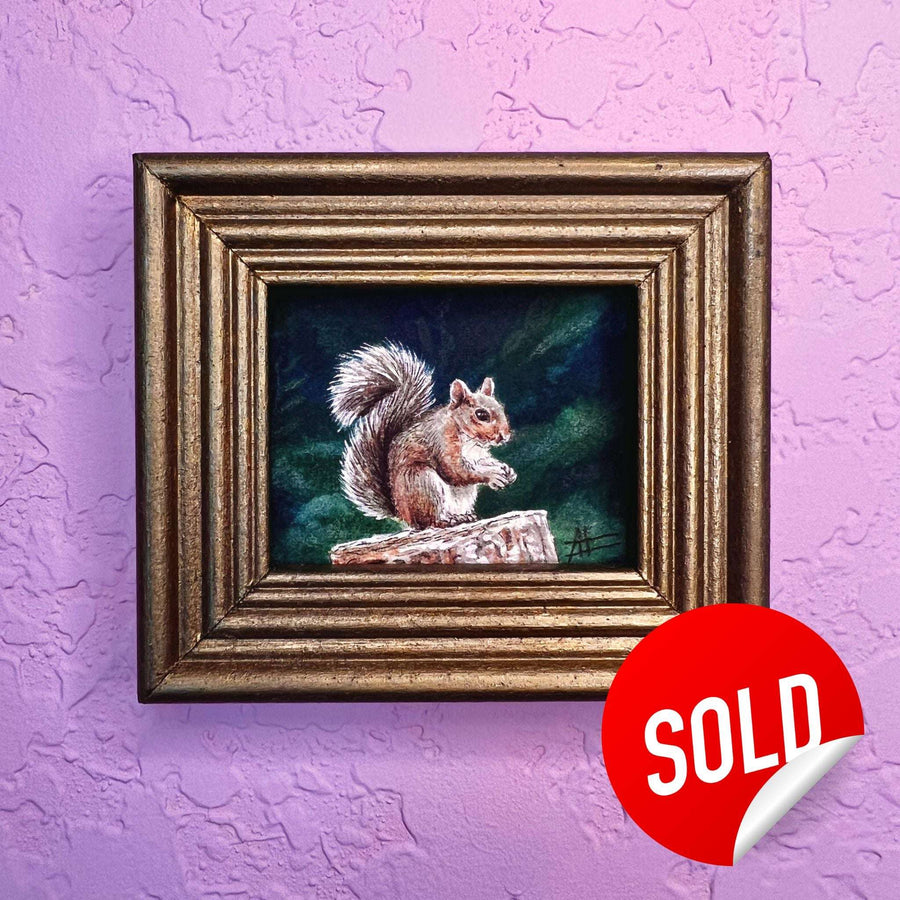 Tiny framed watercolor painting of a squirrel on a log with a "SOLD" sticker on a purple textured wall.