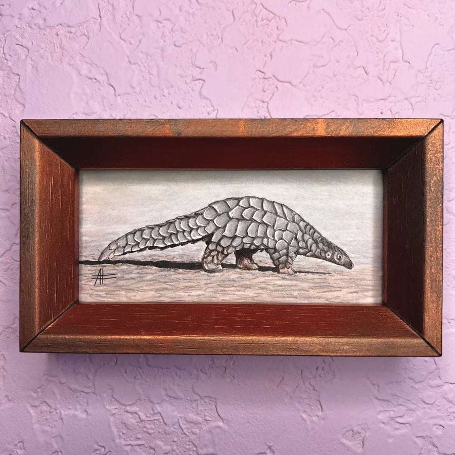 Richly framed miniature watercolor painting of a pangolin on a muted backdrop, placed on a purple wall.