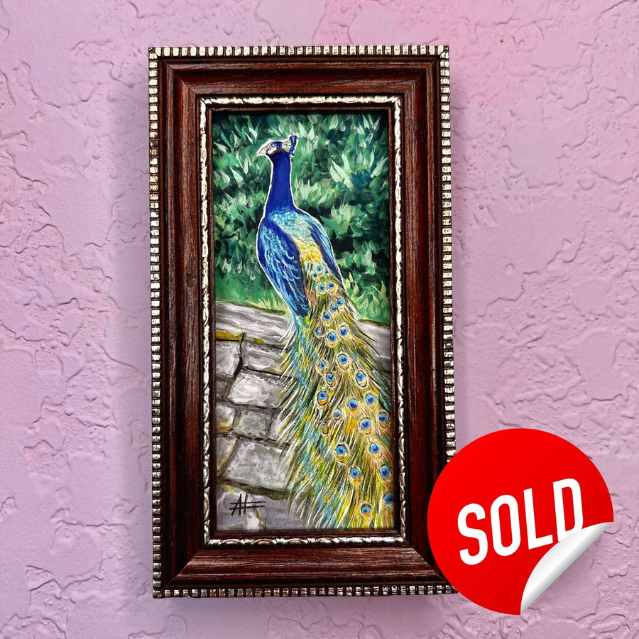 Vibrant miniature peacock painting with its plumage on display in a dark wooden frame, marked sold on a purple wall.