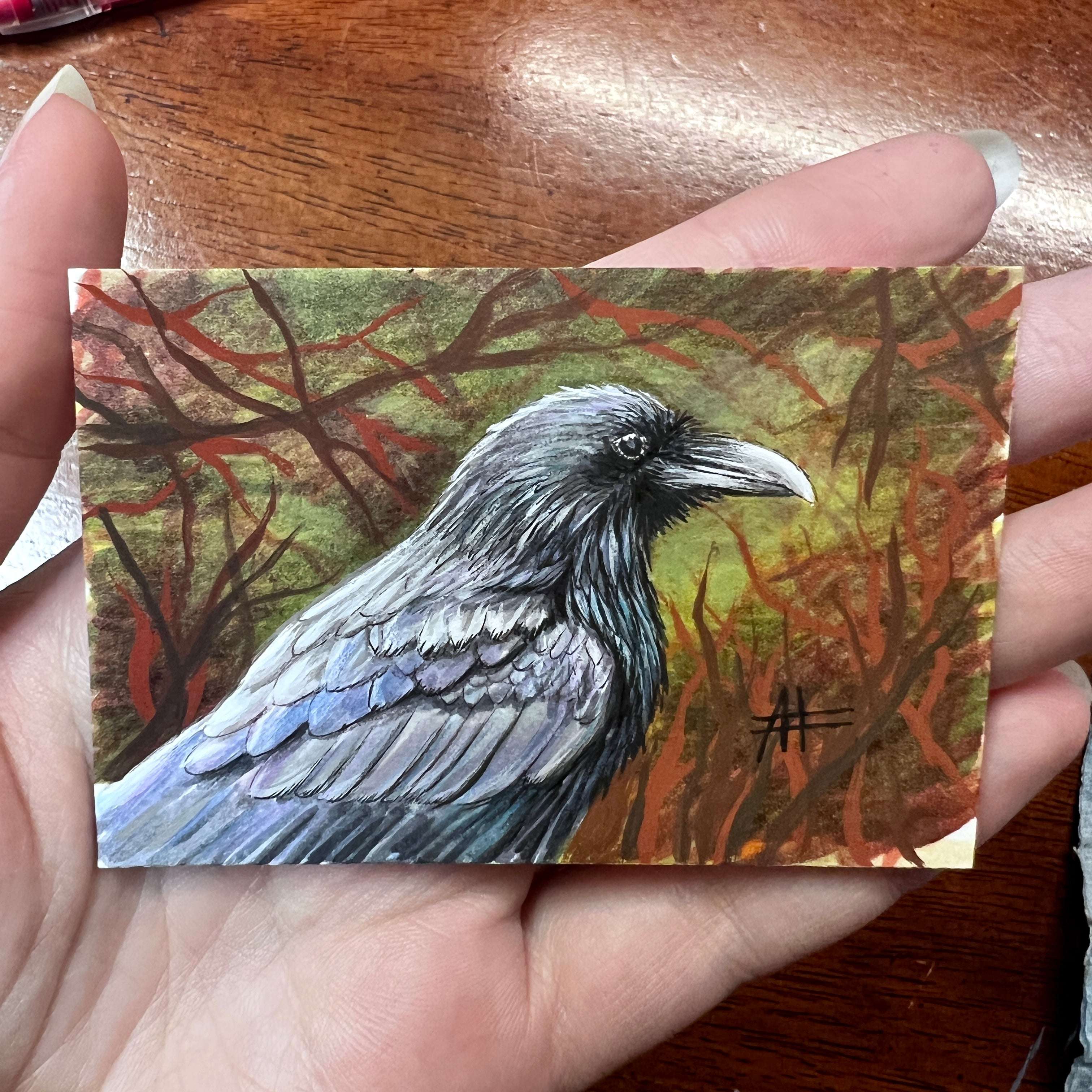 Hand holding a small raven painting with a rich, tangled forest background.