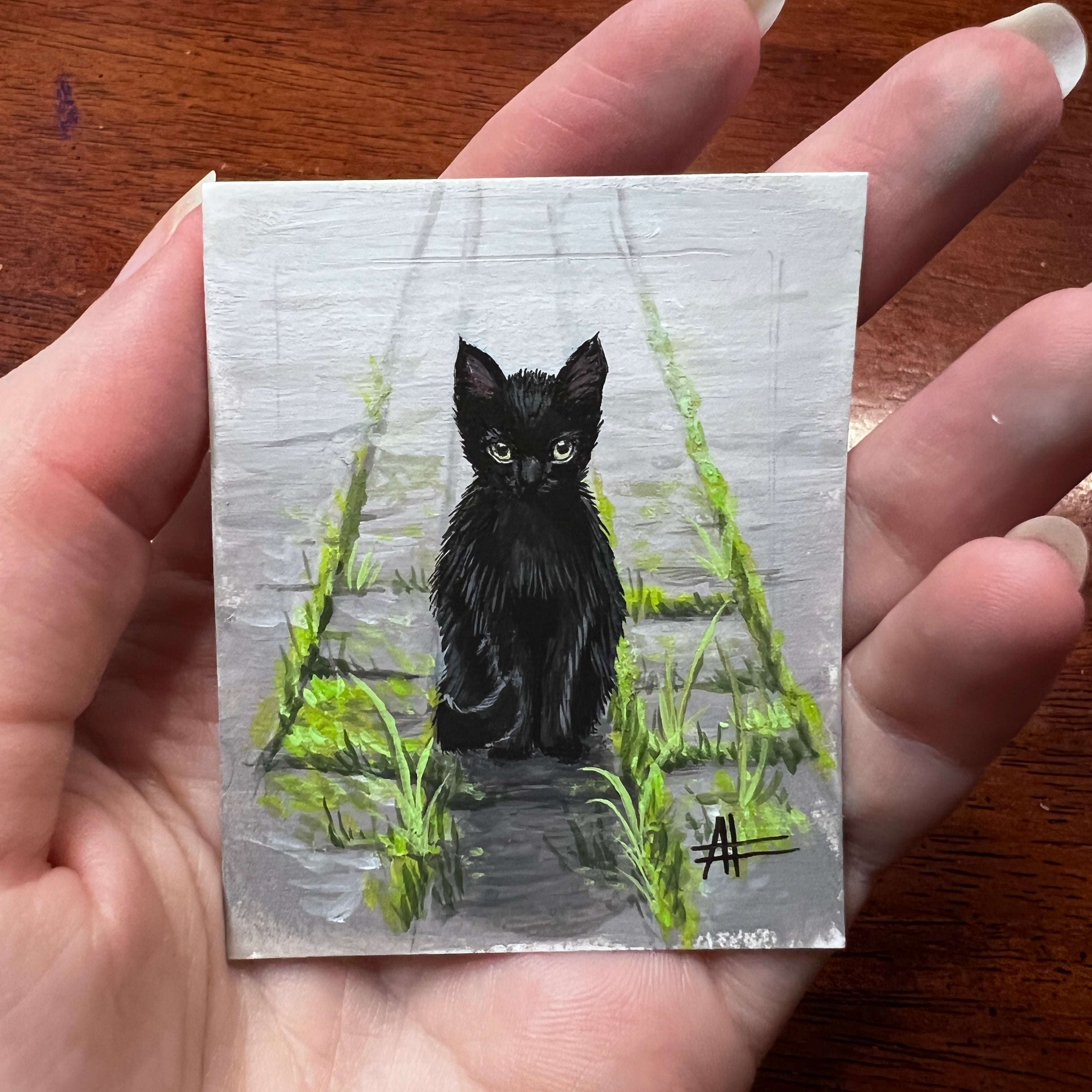 Hand-held painting of a black cat sitting on stone steps among greenery.