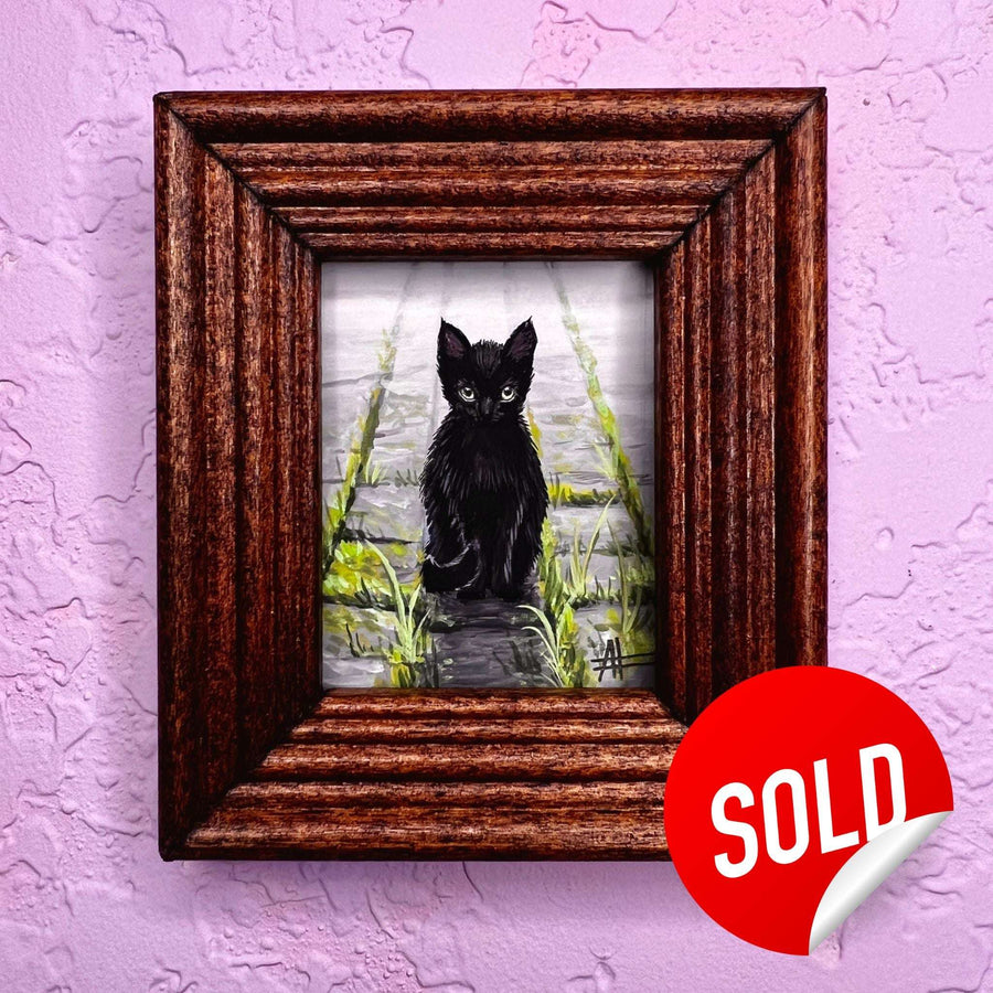 Black cat painting in a dark wood frame on a purple wall, with a red 'SOLD' sticker.