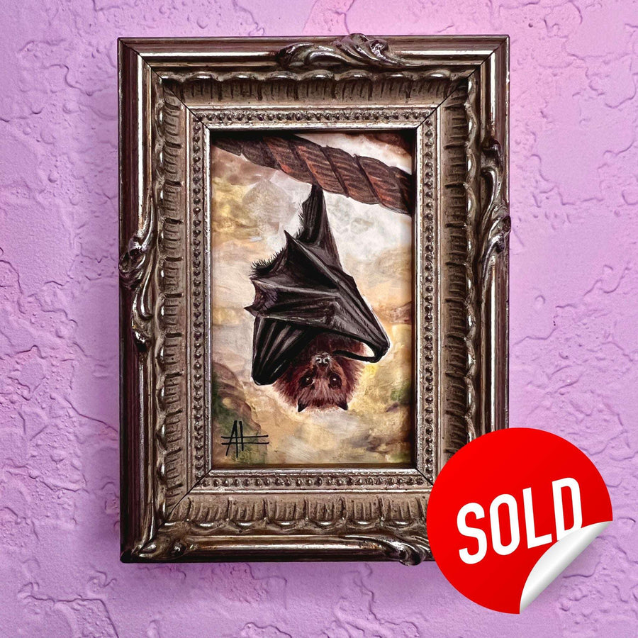 Hanging bat painting with a vivid backdrop in an ornate frame marked as sold on a purple wall.