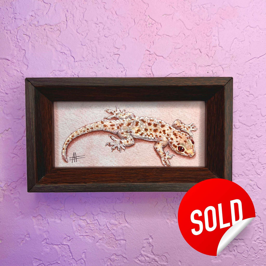 A realistic painting of a gecko with mottled brown and white spots, displayed in a dark wooden frame marked sold on a purple wall.