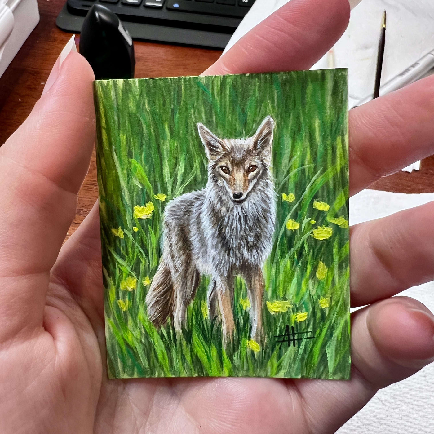 A hand holding a small painting of a coyote amidst yellow flowers with art supplies in the background.