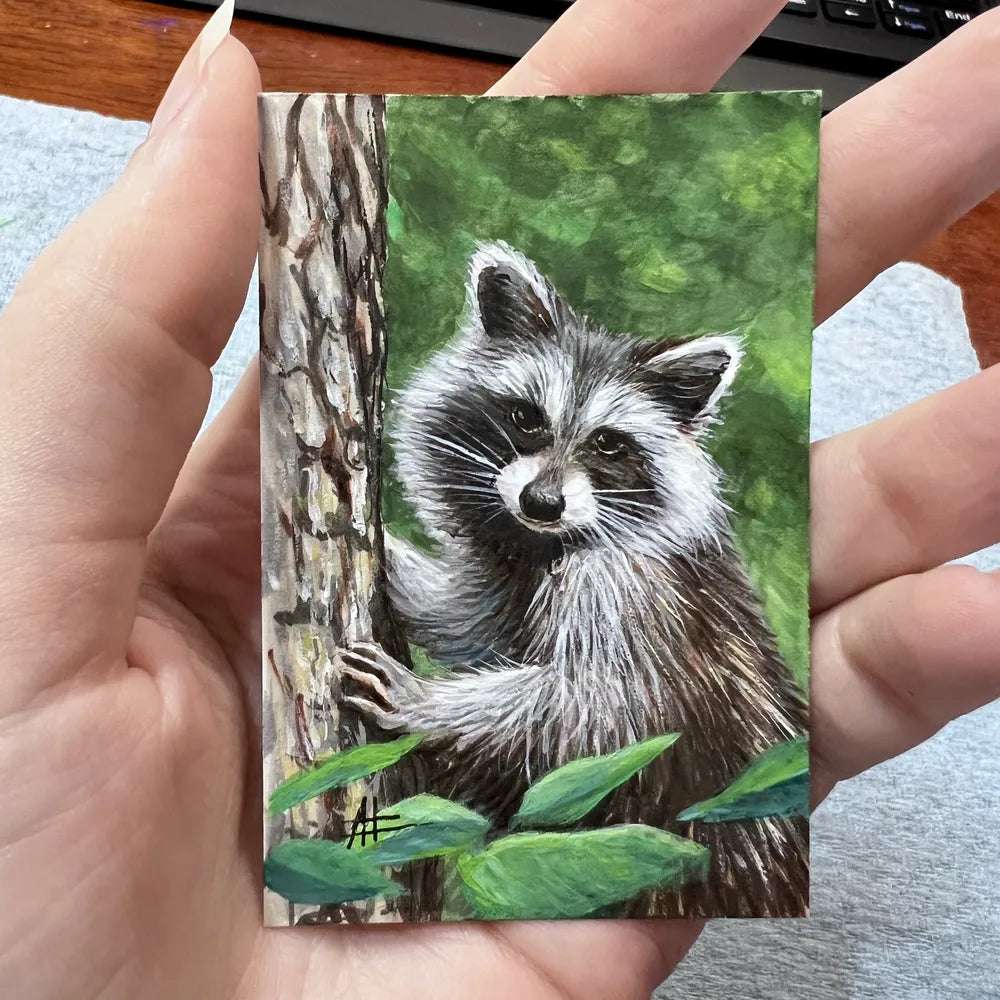 Small raccoon painting held between fingers with a keyboard in the background.