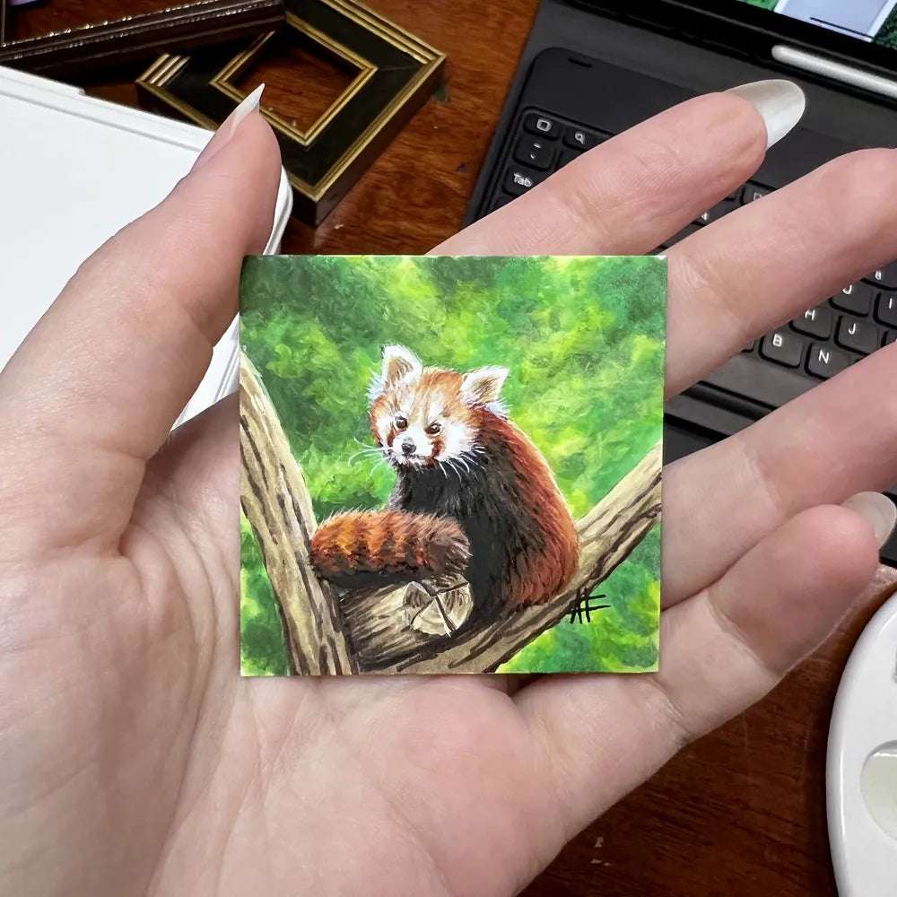 Miniature painting of a red panda on a branch held in hand with art supplies in the background