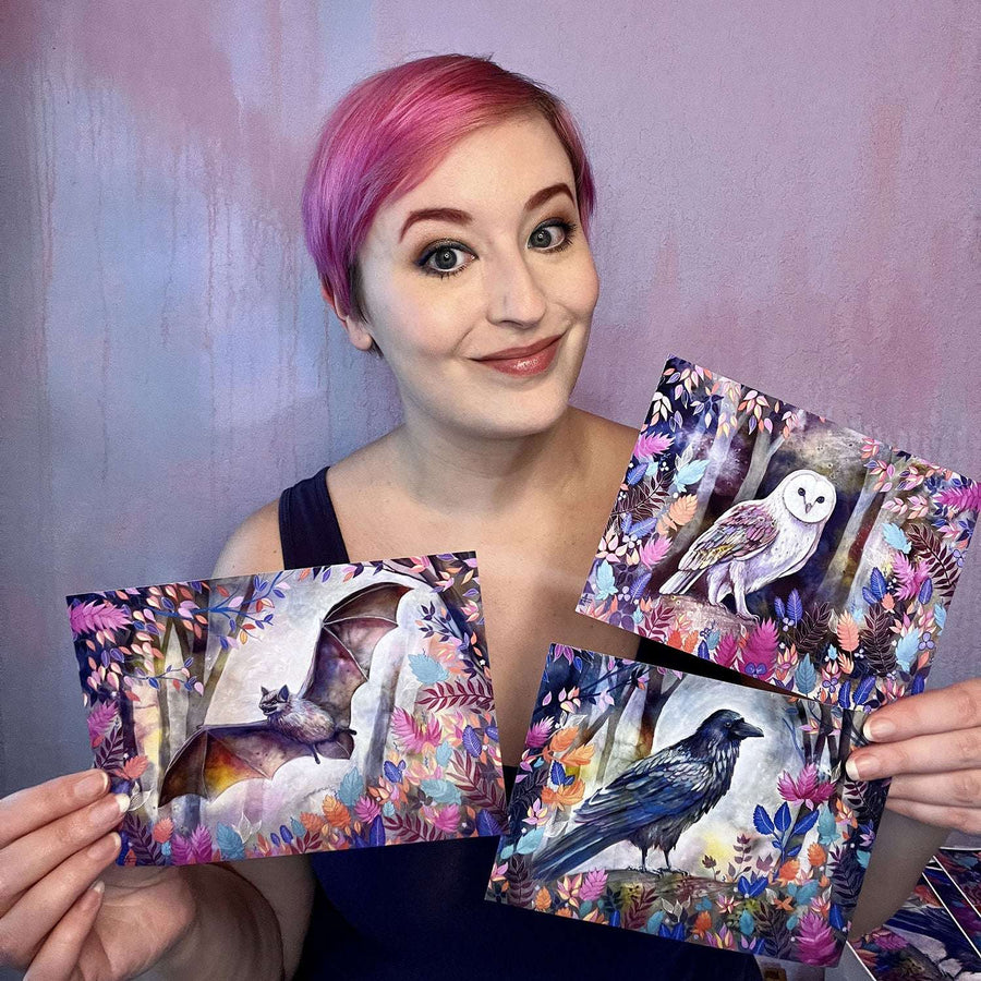 Smiling woman presenting three small prints of owl, bat, and raven in a colorful forest setting.