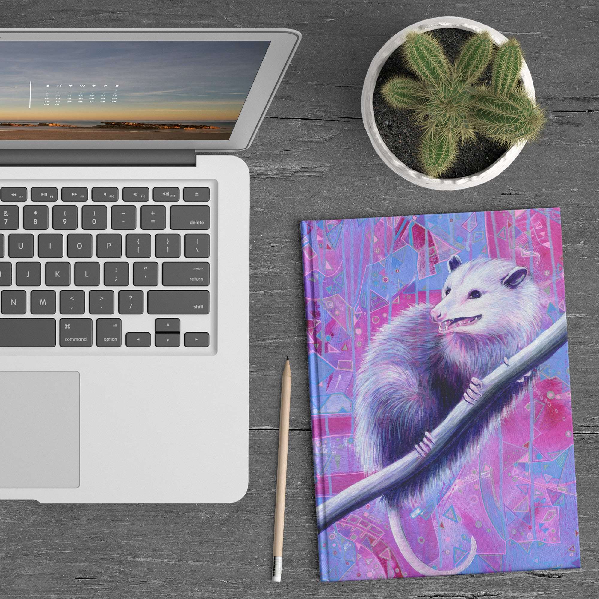 A laptop next to an Opossum Journal with a vibrant illustration of a possum on a colorful geometric background, a pencil, and a small potted cactus on a wooden desk.