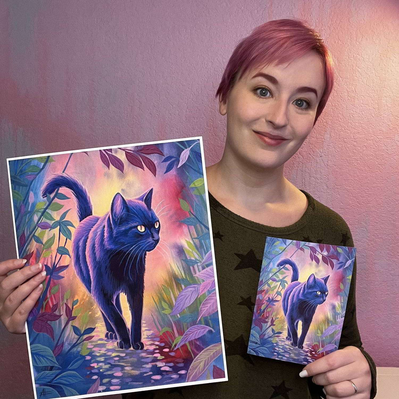 A woman with short pink hair smiles while holding up a large and a small print of "The Cat (Twilight Watch)" featuring a black cat in a colorful forest.