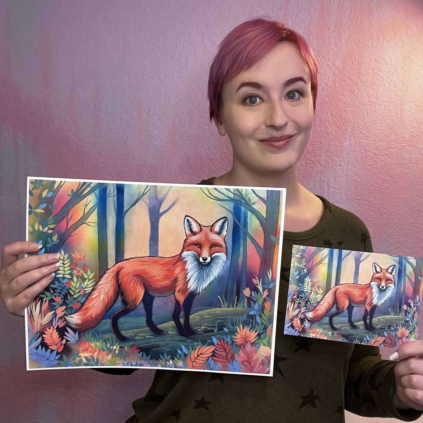 Smiling woman with pink hair presenting two differently sized art prints depicting a red fox in a mystical twilight forest.
