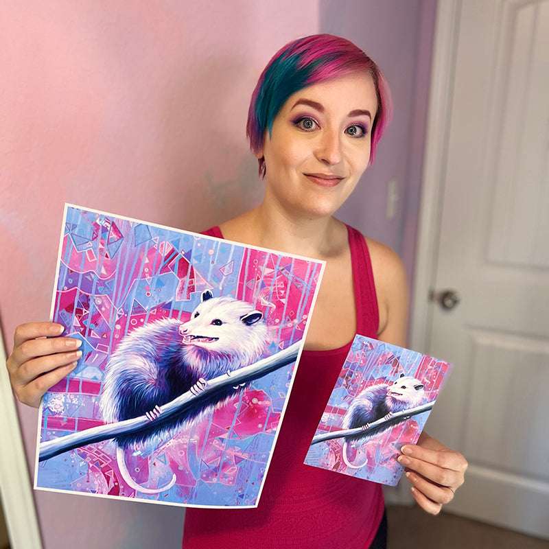 Smiling woman with pink and blue hair holds art prints of a white opossum on a branch with vivid abstract designs.
