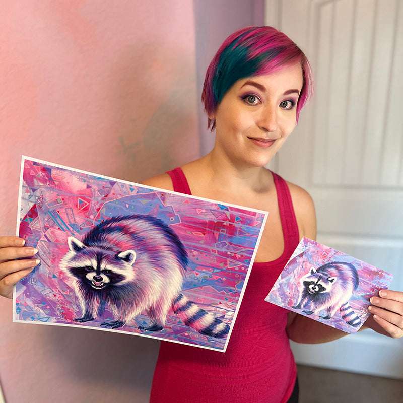 A woman with short, multi-colored hair smiles while holding two colorful prints of The Raccoon (Trash Animals) - Fine Art Print against a pink wall background.