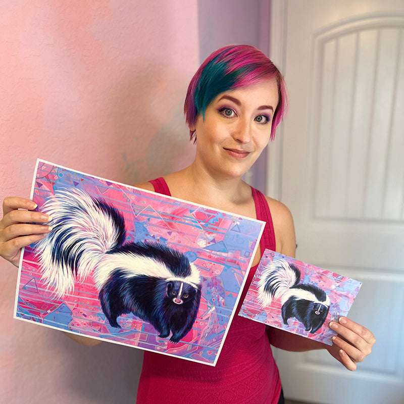 Woman with vibrant hair smiling and holding two art prints of a skunk against a complex pink and blue abstract background.