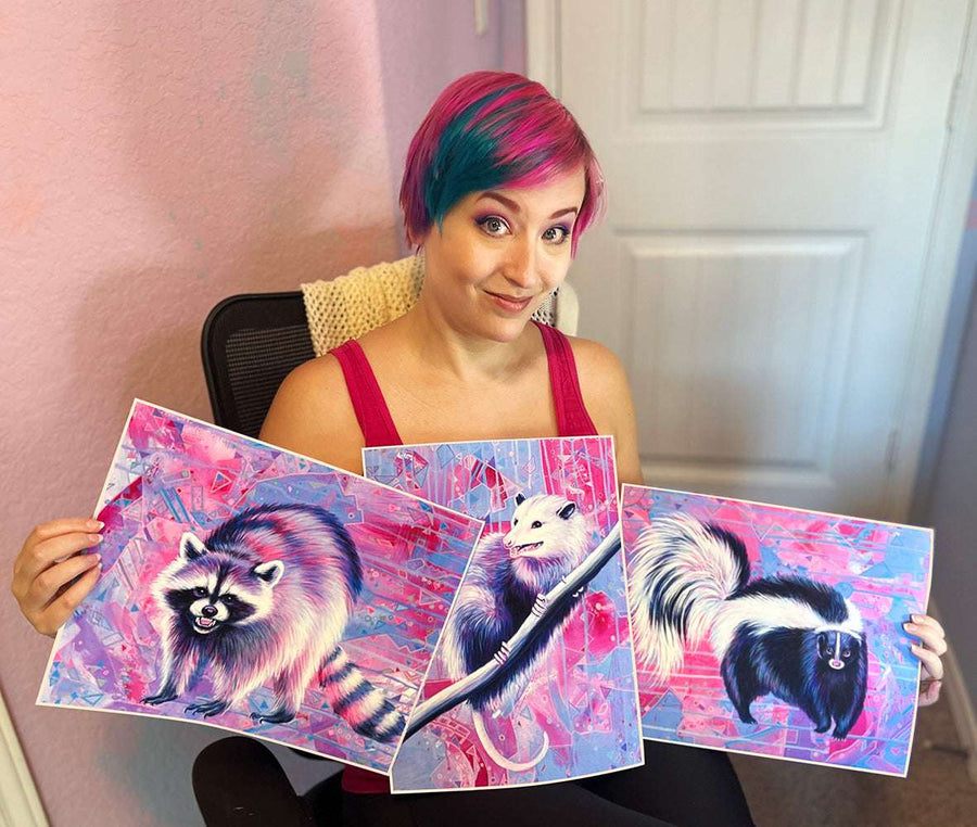 A woman with pink hair displays three colorful paintings of raccoons from the Trash Animals - Fine Art Print Bundle in a home setting.