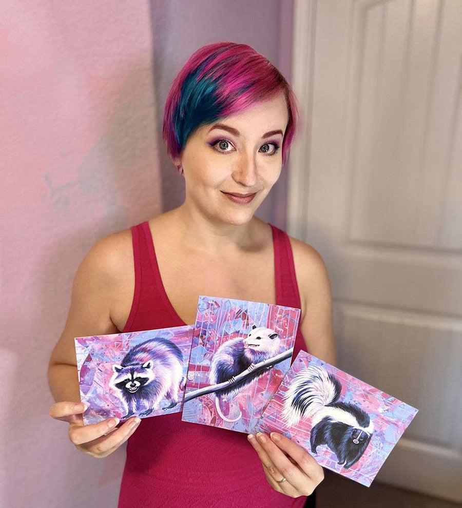 A woman with colorful hair holding three Trash Animals - Fine Art Print Bundle of skunks in a pink-themed room.