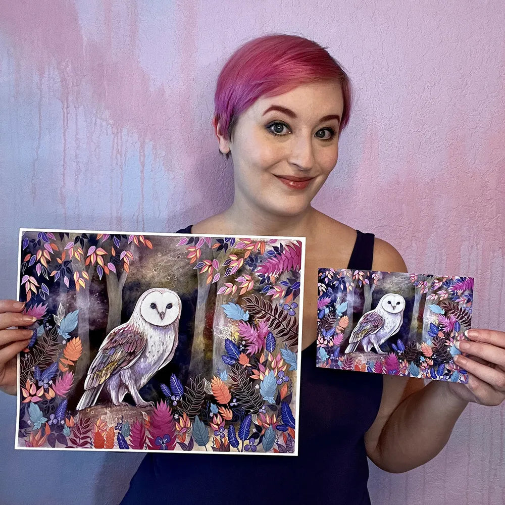 A woman with pink hair smiling and holding two paintings of The Owl (Night Flight) amid colorful foliage.