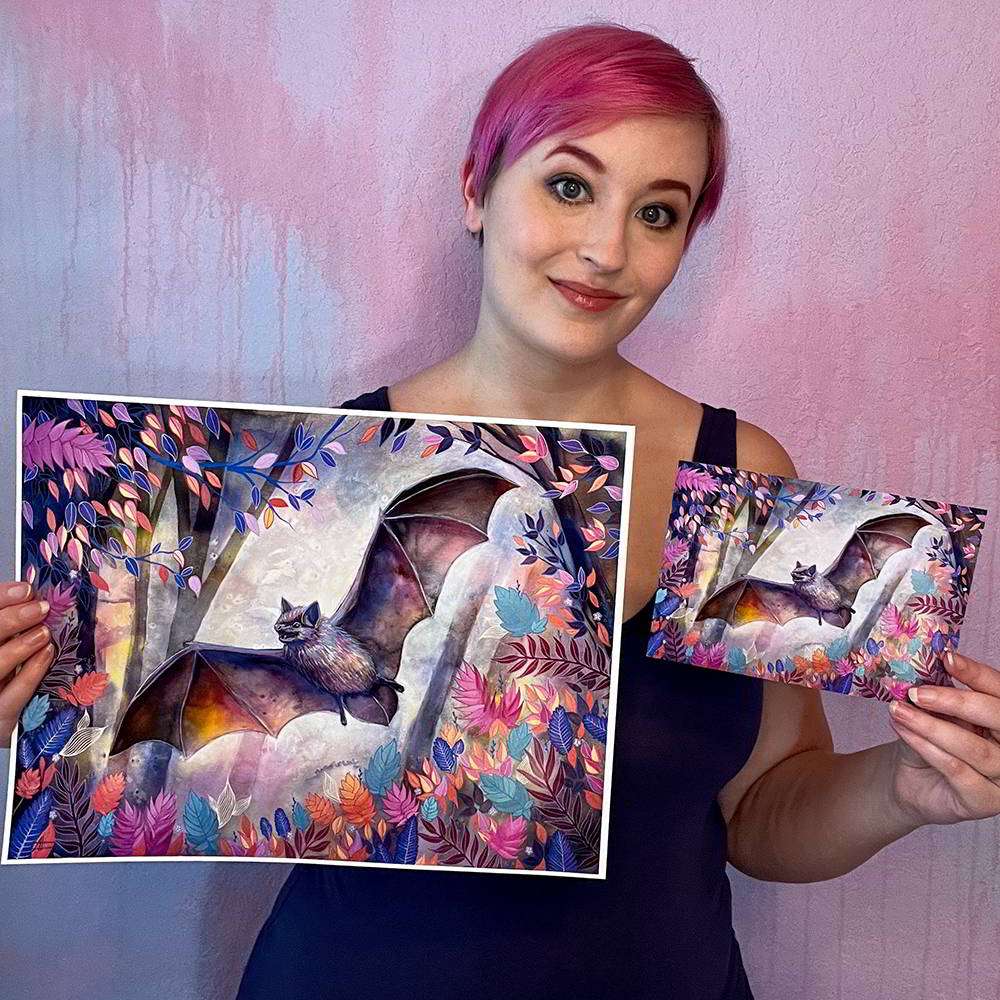 A woman with pink hair holds two "The Bat (Night Flight) - Fine Art Print" featuring vibrant, whimsical designs with a bat subject and foliage motifs against a purple wall.