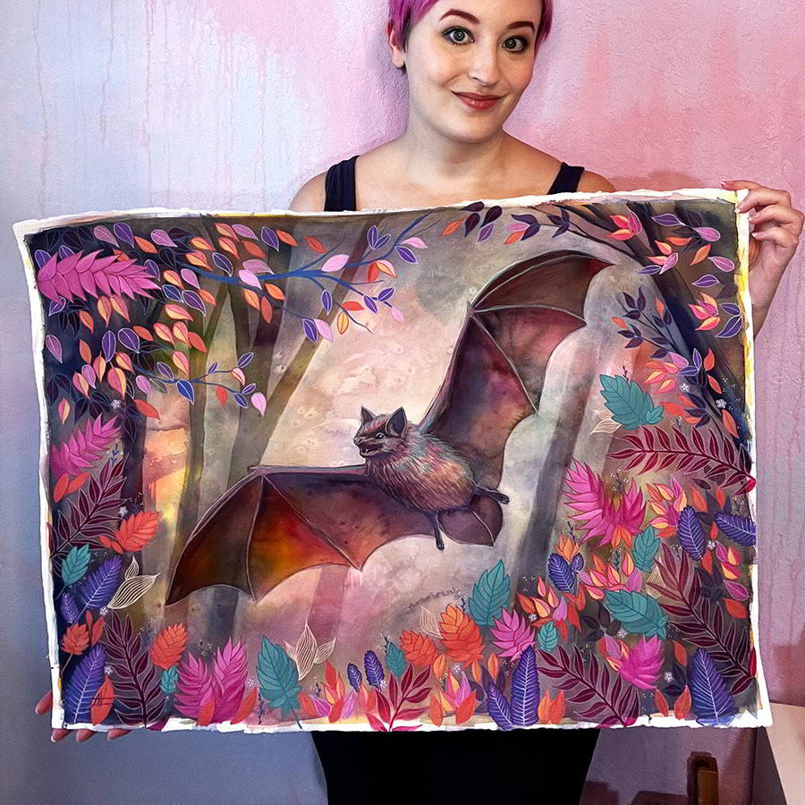 A woman smiling and holding an original watercolor artwork titled "The Bat (Night Flight)", depicting an in-flight bat surrounded by vibrant foliage.