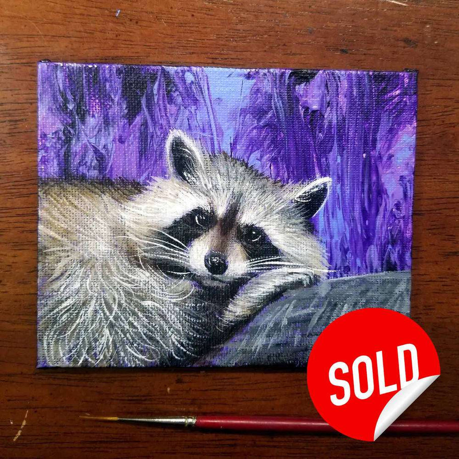 A small painting of a Tiny Raccoon peeking from behind a log against a purple background, marked with a red "sold" sticker.